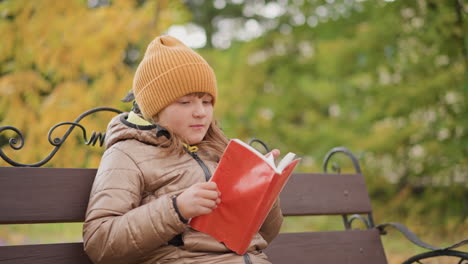 little girl reading red cover book on park bench surrounded by golden autumn leaves glancing up at swaying branches with curious smile capturing tranquil outdoor reading and seasonal