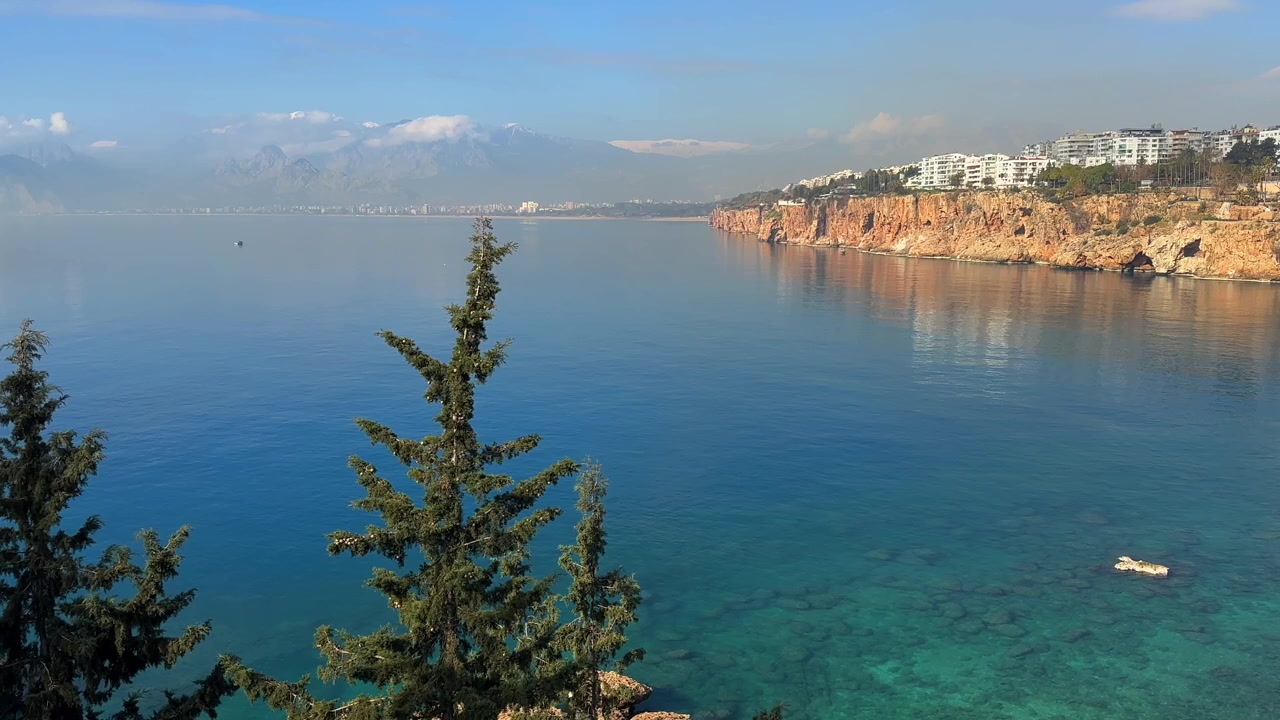 Scenica costa di Konyaalti ad Antalya, caratterizzata da scogliere rocciose, acqua turchese del Mediterraneo, le distanti Montagne del Tauro e un paesaggio costiero pacifico sotto un cielo sereno.
