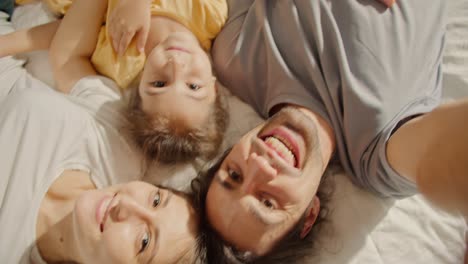 Close-up-shot-of-a-brunette-man-in-a-gray-T-shirt-taking-a-selfie-with-his-wife-a-brunette-girl-and-their-daughter-a-little-brunette-girl-in-a-yellow-dress-in-a-family-lying-on-the-sofa-and-taking-a-selfie-from-above-in-a-cozy-room