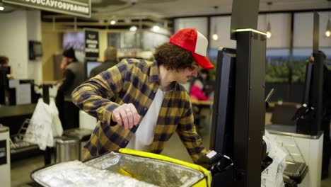 Confident-food-delivery-guy-in-a-plaid-shirt-punches-goods-at-the-checkout-and-then-puts-them-in-his-yellow-bag-for-food-delivery-in-the-supermarket