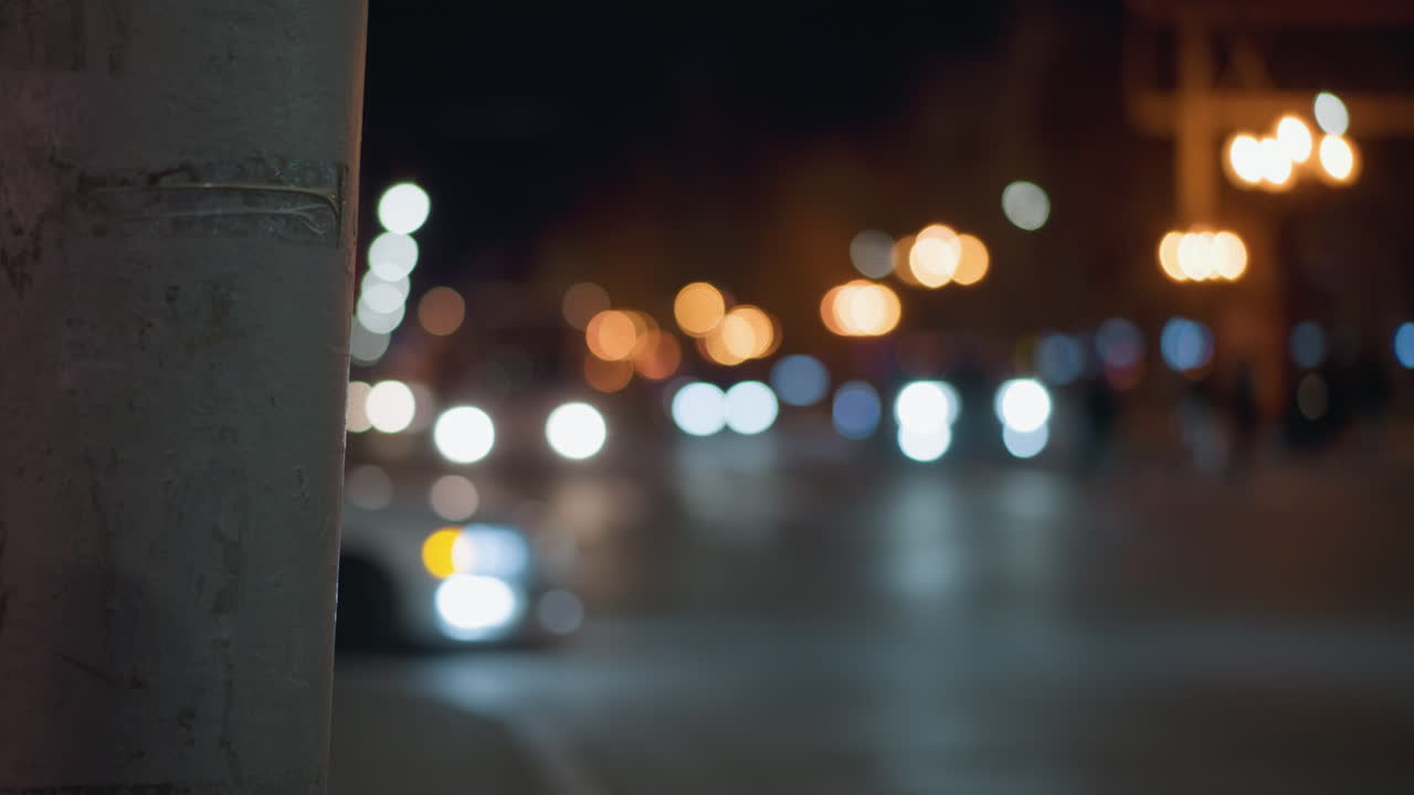 vista bokeh de una escena nocturna de la ciudad con coches borrosos y luces de la calle en el fondo, capturada con un enfoque en un poste de la calle cercana