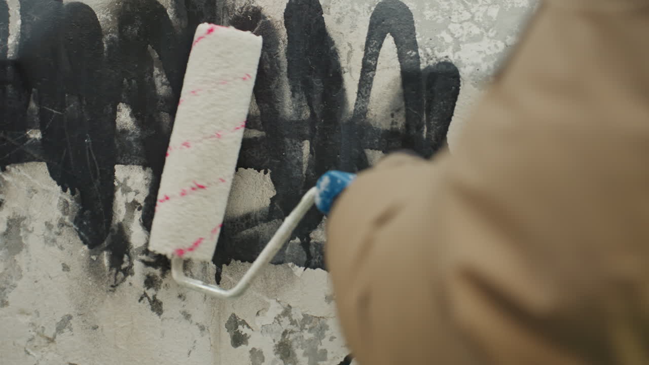Wide shot painter covering black graffiti on aging concrete facade, volunteer worker in brown jacket rolling paint to remove tags, visible streaks and drips, methodical coating and touchup, community