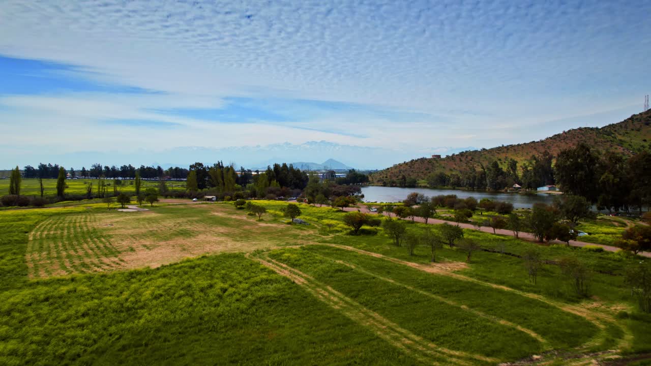 Aerial view dolly in from a meadow of the Caren lagoon park towards the Plomo hill with a commercial airplane taking off, sky with clouds in patterns, Santiago, Chile.