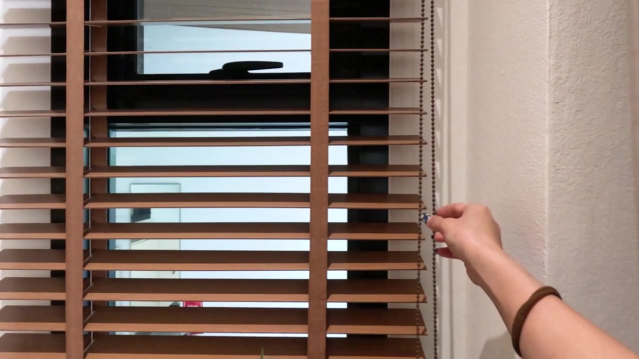 A hand pulls a cord to adjust wooden window blinds beside soft white sheer curtains