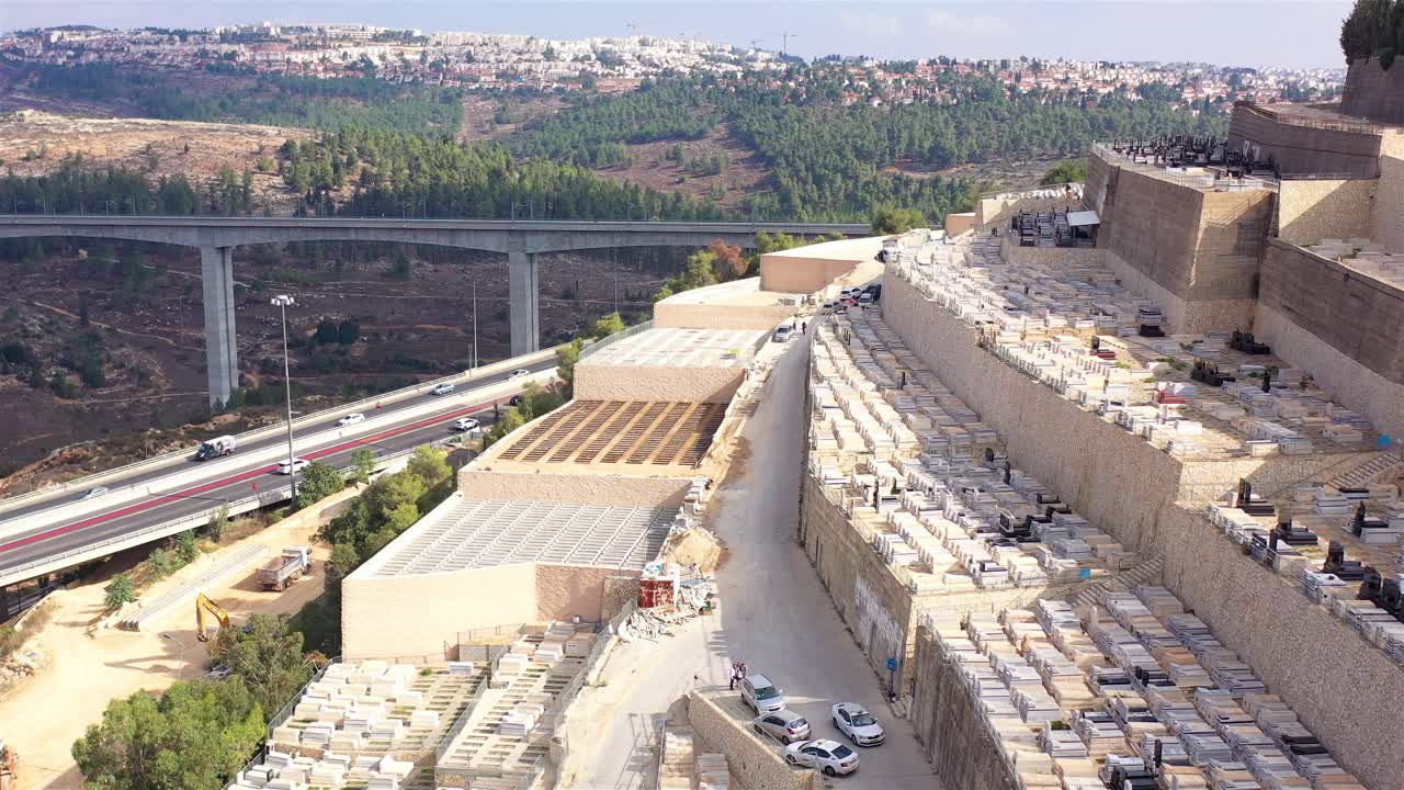 Aerial view of a large terraced cemetery adjacent to a highway bridge, with a city skyline in the distance