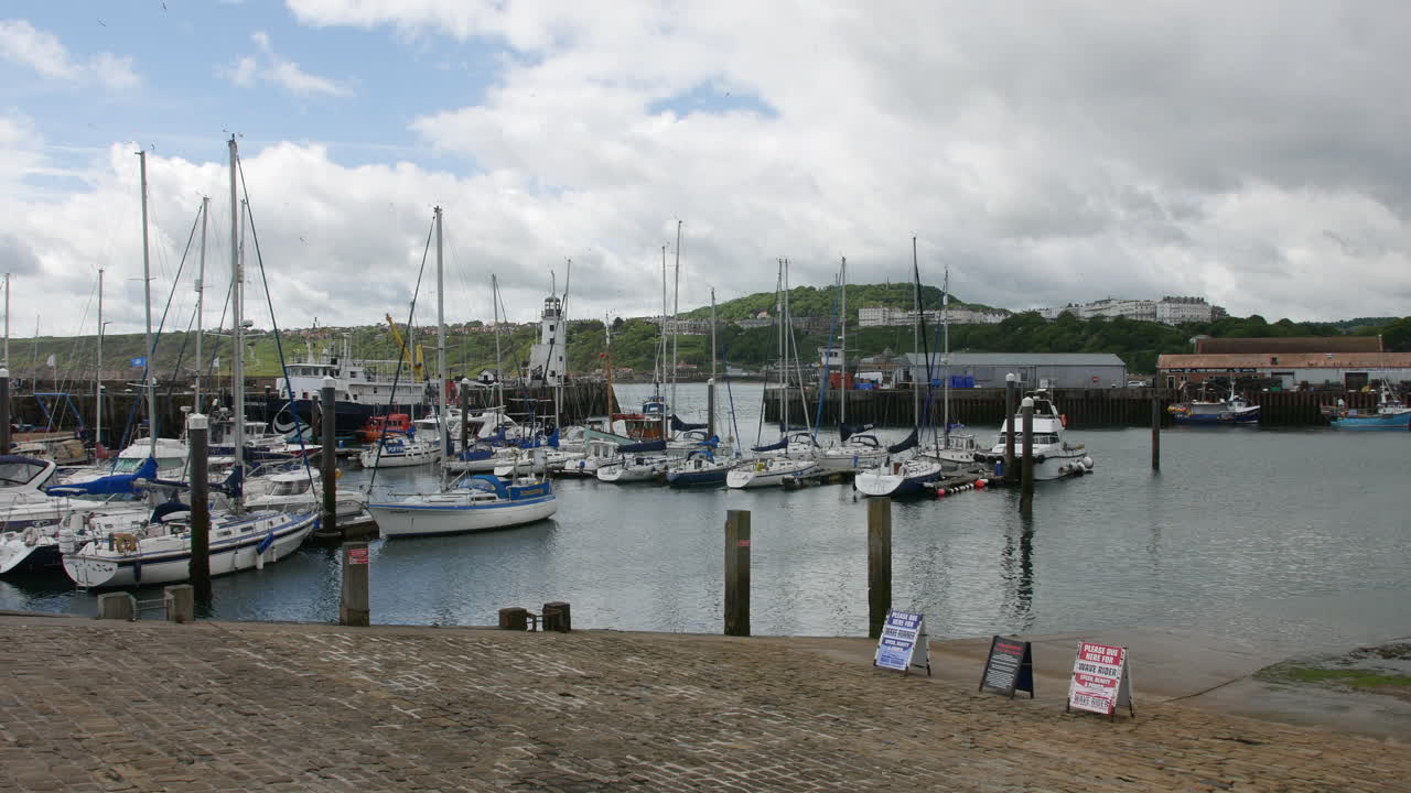 A harbor filled with sailboats and motorboats docked near a lighthouse in Scarborough, North Yorkshire in England, with coastal hills and buildings in the distance