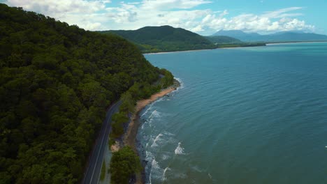 aerial drone bird view of modern silver car driving on road along the seaside coast with sandy beach and lush green tree forest and blue sea waves in 4k