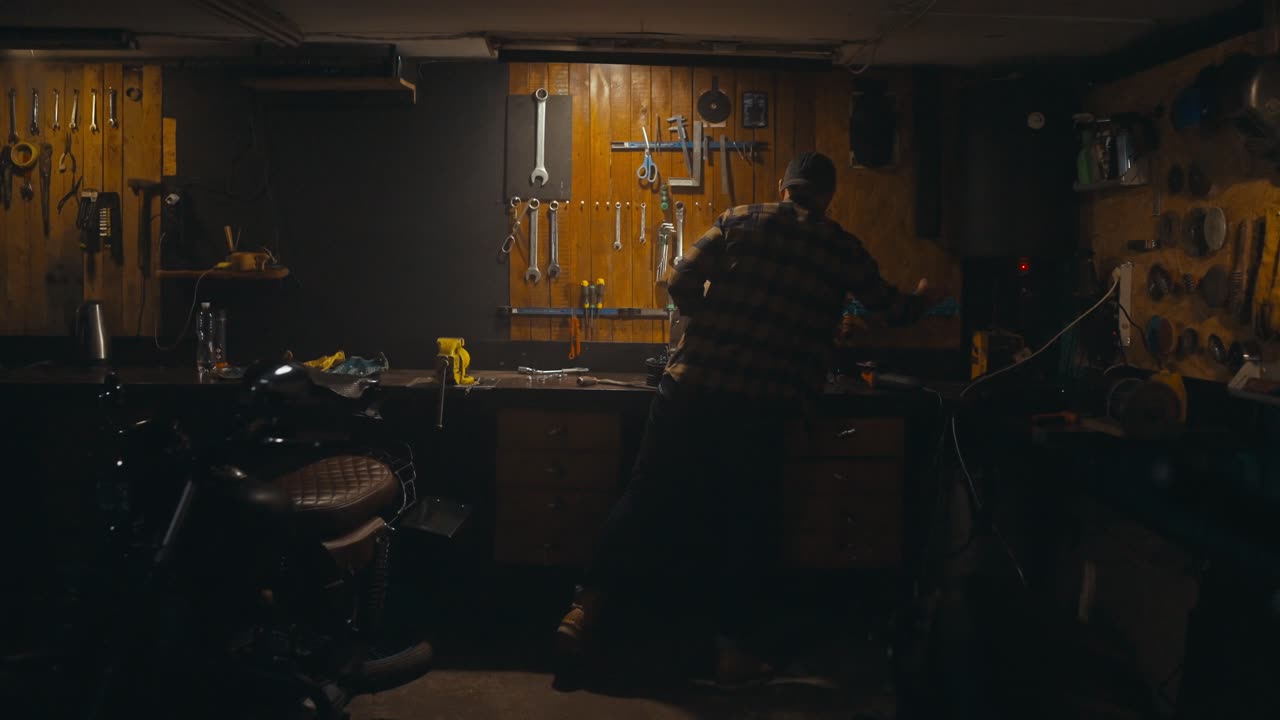 Rear view of a confident guy mechanic in a plaid shirt and yellow gloves working on a workbench among many tools in his cozy studio workshop