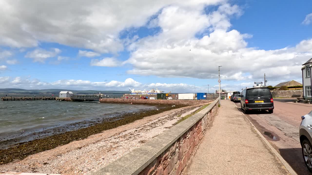 A steady camera walk along Cromarty harbor’s stone seawall, passing parked cars and waterfront buildings under bright daylight with scattered clouds