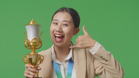 close up of asian business woman in a suit with a gold medal and trophy showing call me gesture and smiling to camera as the first winner on green screen background in the studio