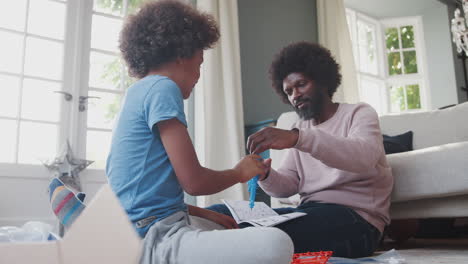 middle aged black father sitting on the floor at home helping his pre teen son with the instructions for a toy construction kit, close up, low angle