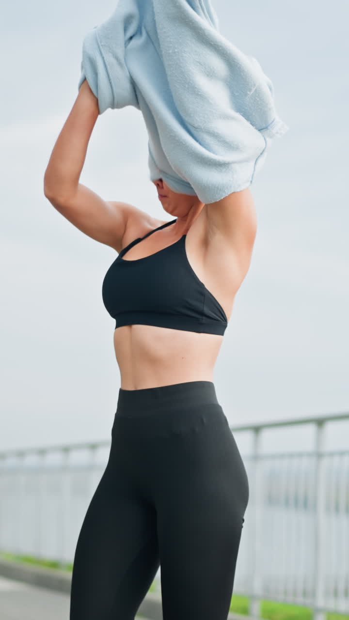 Woman standing in front of iron fence, removing her sweater under sunny weather, ideal for fitness, outdoor, and lifestyle moments in a bright, active environment