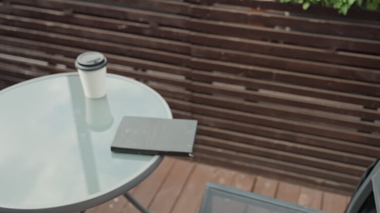 Giovane donna bianca su un balcone che si tiene la testa, giacca bagnata per la pioggerellina, cuffie intorno al collo, smartphone e notebook chiuso sul tavolo di vetro accanto a un bicchiere di carta, espressione pensierosa, si ferma dopo una telefonata tesa