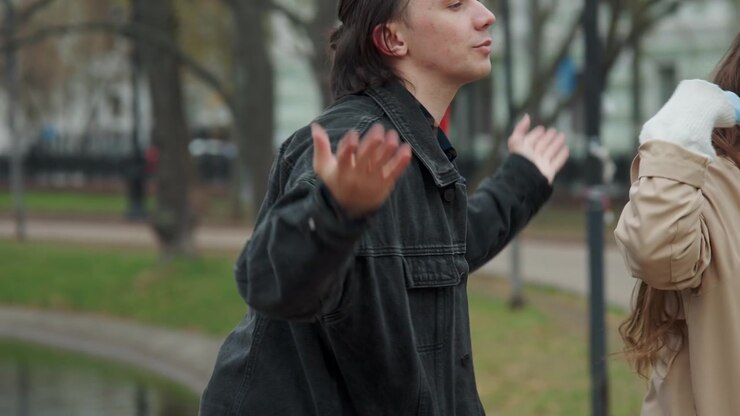 Close up of man and woman interacting outdoors in park setting with gestures indicating conversation or explanation. Man wears dark jacket over plaid shirt while gesturing expressively