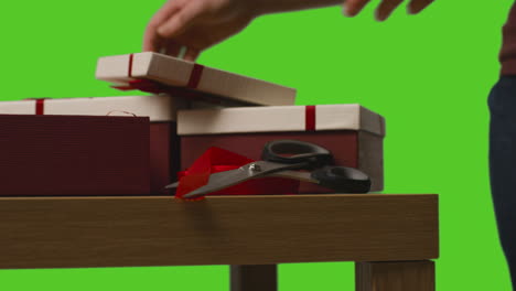 close up of man gift wrapping presents in boxes decorated with ribbon on table shot against green screen 3