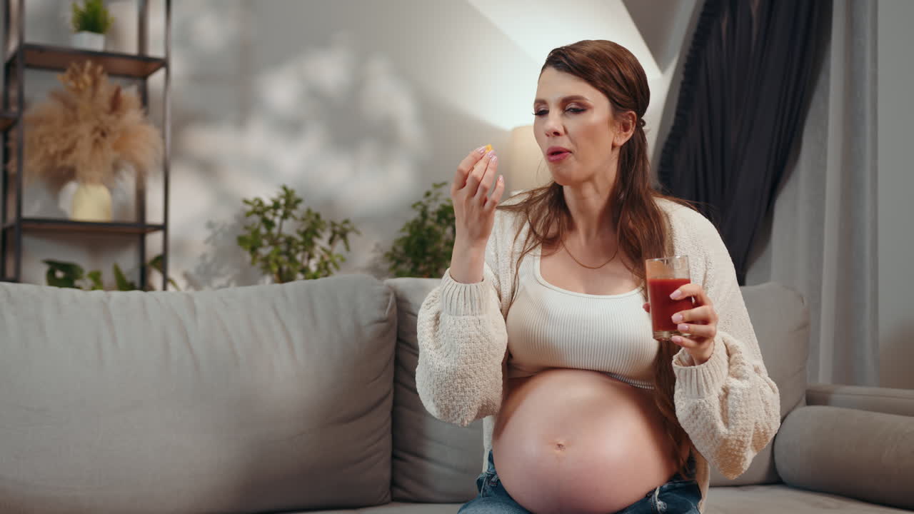 Pregnant woman sits on sofa holding glass of juice and eating fresh fruit for healthy nutrition