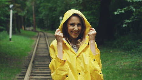 Retrato-De-Una-Mujer-Con-Un-Abrigo-Amarillo-A-Prueba-De-Lluvia-Parada-Frente-A-La-Cámara-Y-Riendo-En-El-Bosque