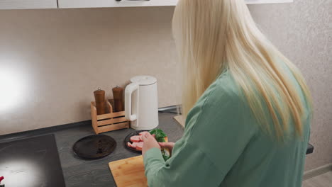 rear view of blonde lady in green shirt cutting open noodles pack on kitchen counter, placing knife down then using hand to tear open pack, near plate of sliced sausage and wooden pepper grinder
