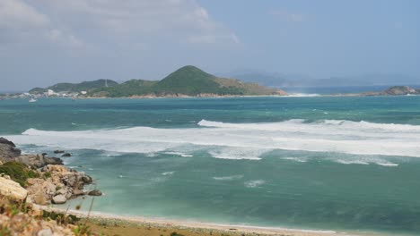 Perfect-waves-washing-up-shore,-green-mountain-island-on-the-sunny-horizon