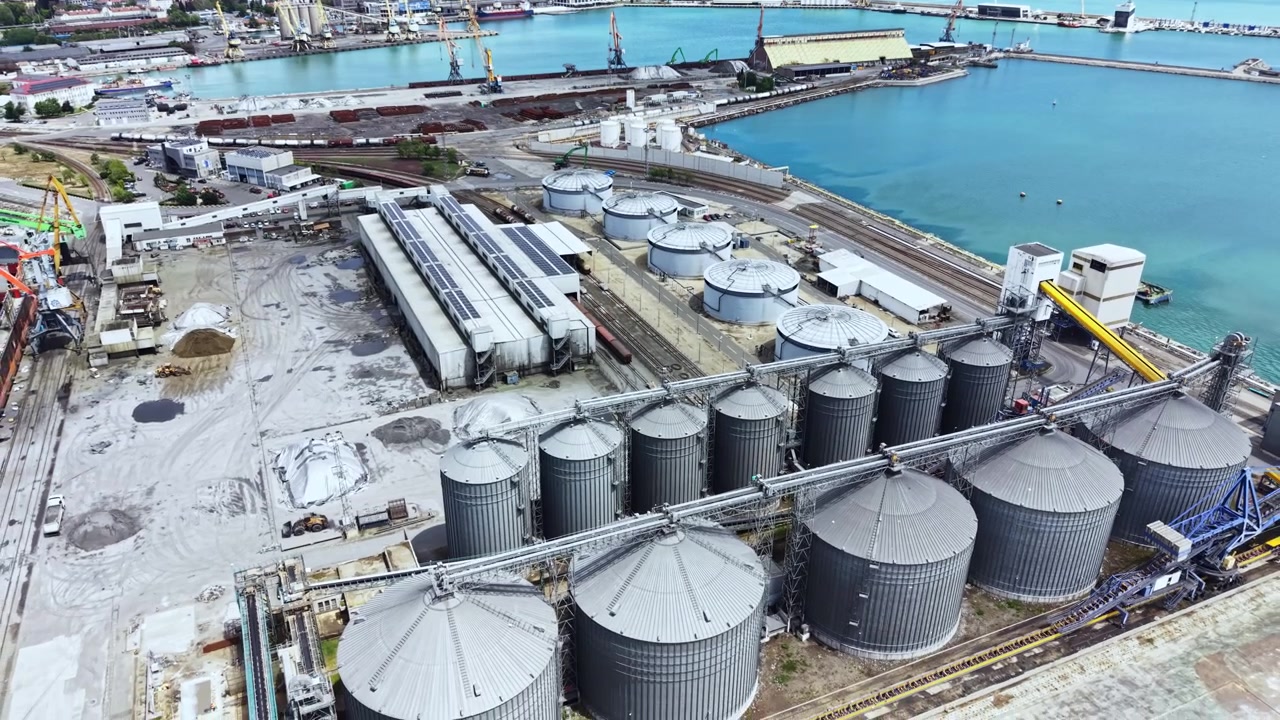 Großes Industriehafengebiet mit Lagersilos, Laderampen und Seecontainern bei Tageslicht in einer Küstenstadt.