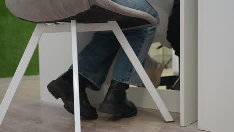 close-up view of customer in jeans and black boots seated on soft chair with white legs during manicure session while technician in mask and pink gloves works attentively out of frame at nail station