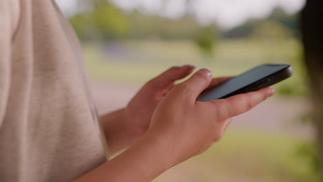 close-up of individual s hand typing on smartphone with focus on hands, set against a naturally blurred green background with unclear figures in the distance