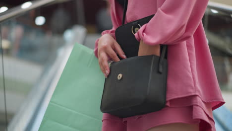 young lady in pink dress and black top, riding an upward-moving escalator, searches through her black bag with concentration and a thoughtful expression, a mint green shopping bag rests by her side