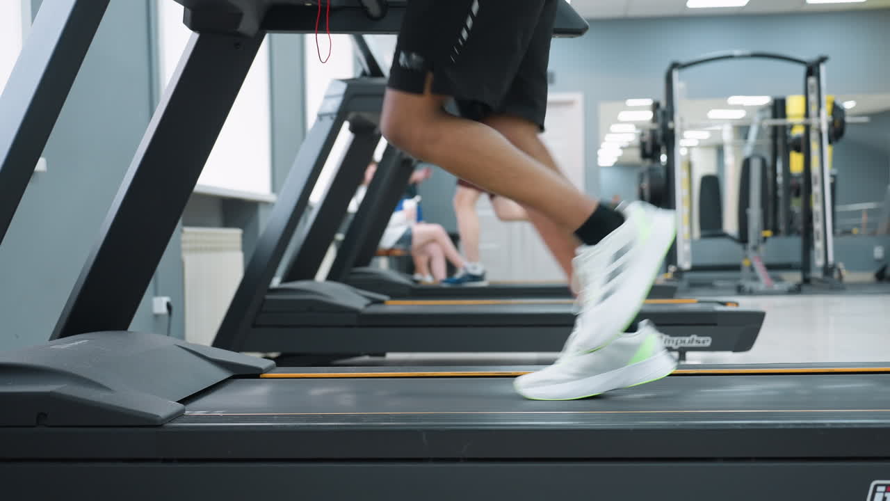 leg view of two people running on treadmills in gym, white sneakers in motion, blurred figures seated in background, gym machines and mirrored wall create depth in bright fitness environment