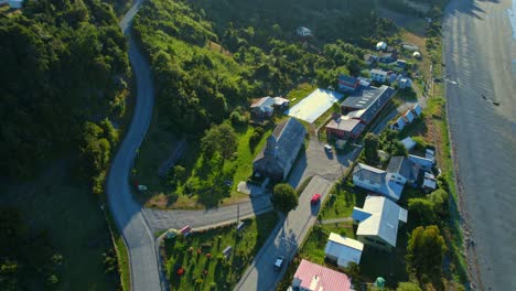 Detif-church,-a-unesco-world-heritage-site-on-chiloé-island,-chile,-with-surrounding-village-and-coastline,-at-golden-hour,-aerial-view