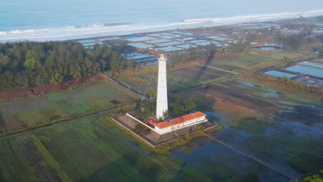 aerial orbit shot of lighthouse near beach and ocean in central java, indonesia - agricultural plantaiton fields in the morning