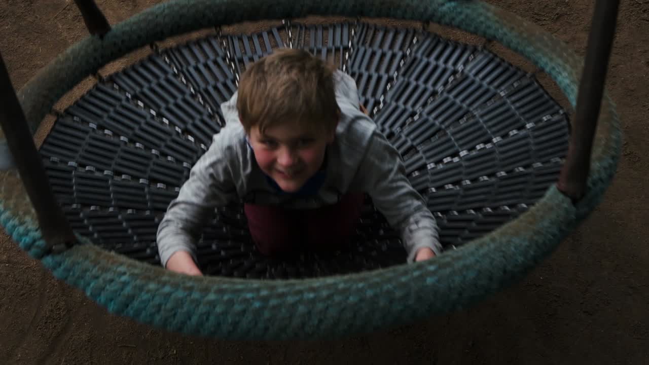 Cheerful carefree young boy sitting in large round park swing activity looking up at camera getting closer