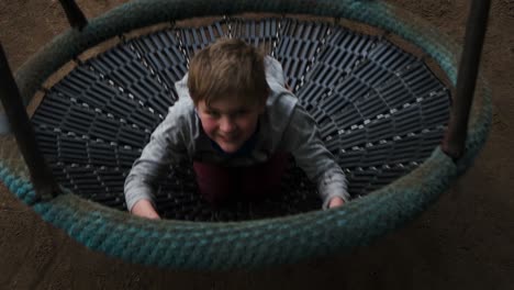 cheerful carefree young boy sitting in large round park swing activity looking up at camera getting closer