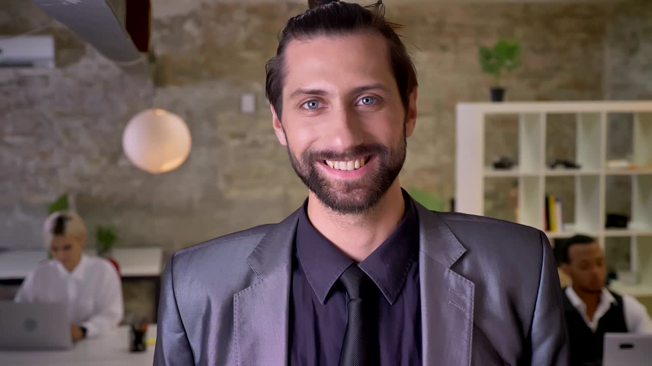apuesto hombre de negocios con barba está mirando a la cámara en la oficina, sonriendo, colegas están trabajando con computadoras portátiles, concepto de trabajo
