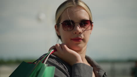 white woman in grey shirt and sunglasses holding shopping bags backward, while standing outdoors with a focused expression, the background is slightly blurred with people partially seen