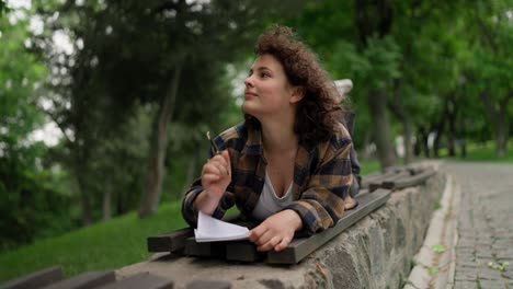 a thoughtful brunette girl with curly hair writes down her ideas in a notebook while lying on a bench in the park