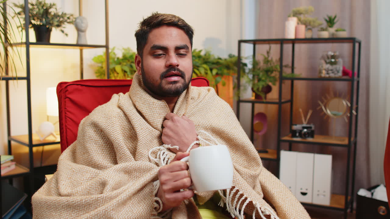 Sick indian man wrapped in plaid sit alone shivering trembling from cold on chair drinking hot tea