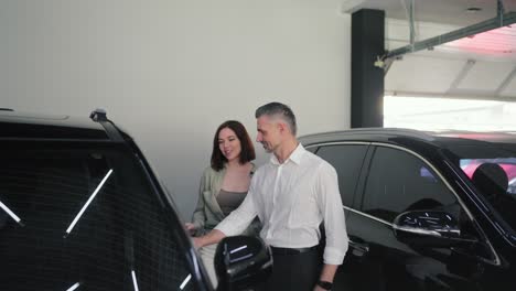 un hombre confiado de mediana edad con una barba gris y con una camisa blanca se está comunicando con su esposa morena y su hijo pequeño sobre un coche en un concesionario de automóviles moderno