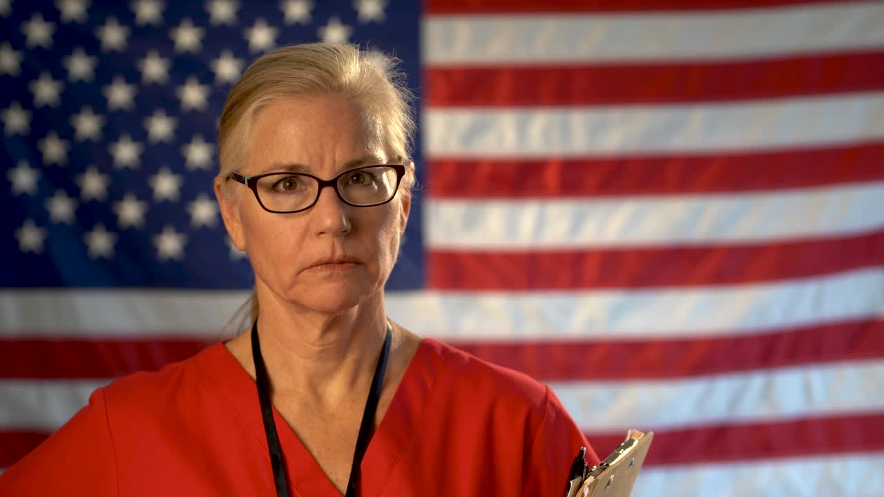 Medium tight portrait of a healthcare nurse with clipboard looking concerned and sad walking towards camera with an out of focus American flag