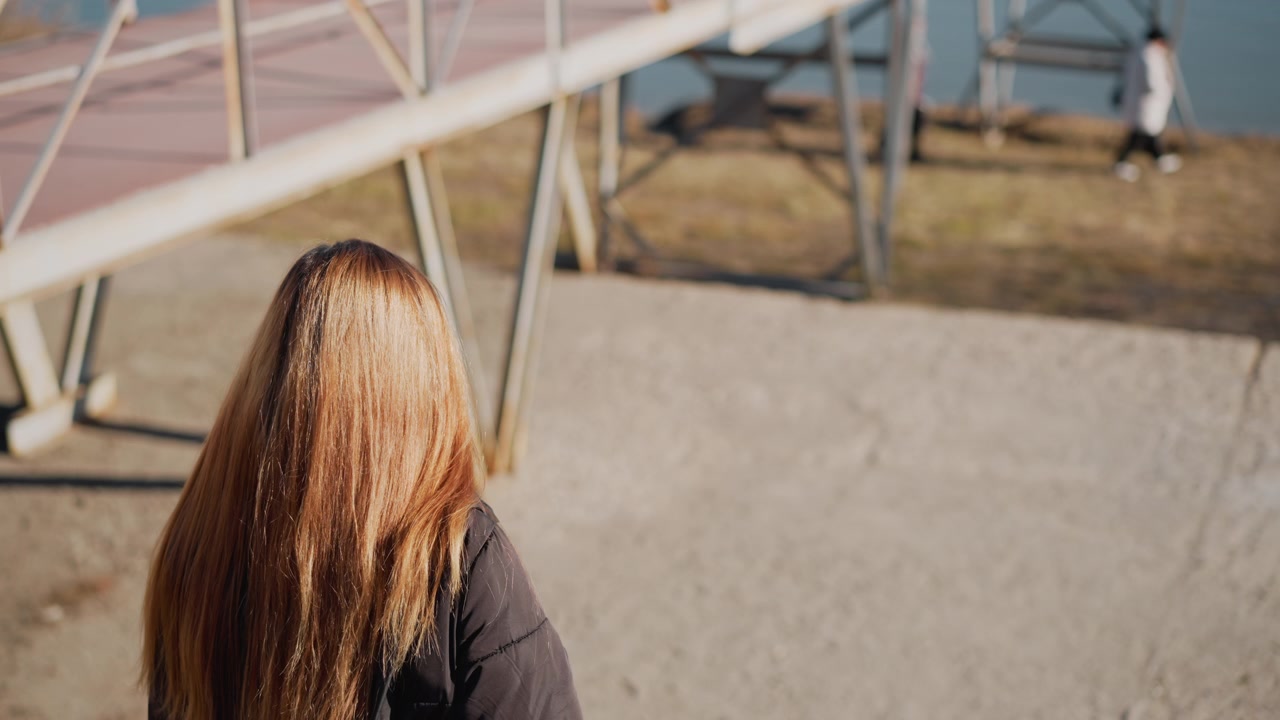 Woman with long hair stands outdoors facing away from camera towards bridge and water. Woman wears dark jacket as people walk in soft daylight on grassy terrain near industrial structures