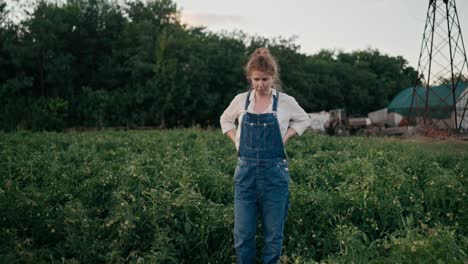 confident girl farmer in denim overalls with red curly hair inspecting plants in a field on a farm