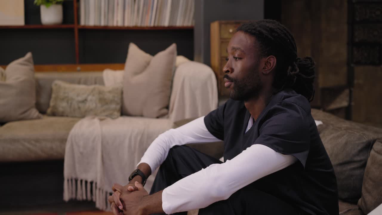 African American man in scrubs entering at home, sitting on floor, talking to camera, copy space