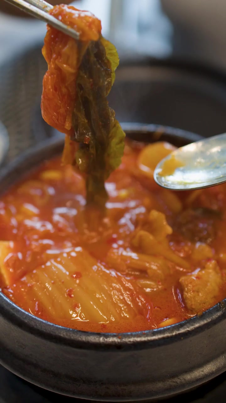 Bidikan close-up sumpit logam mengangkat kubis fermentasi dari sup tahu merah pedas yang mendidih.