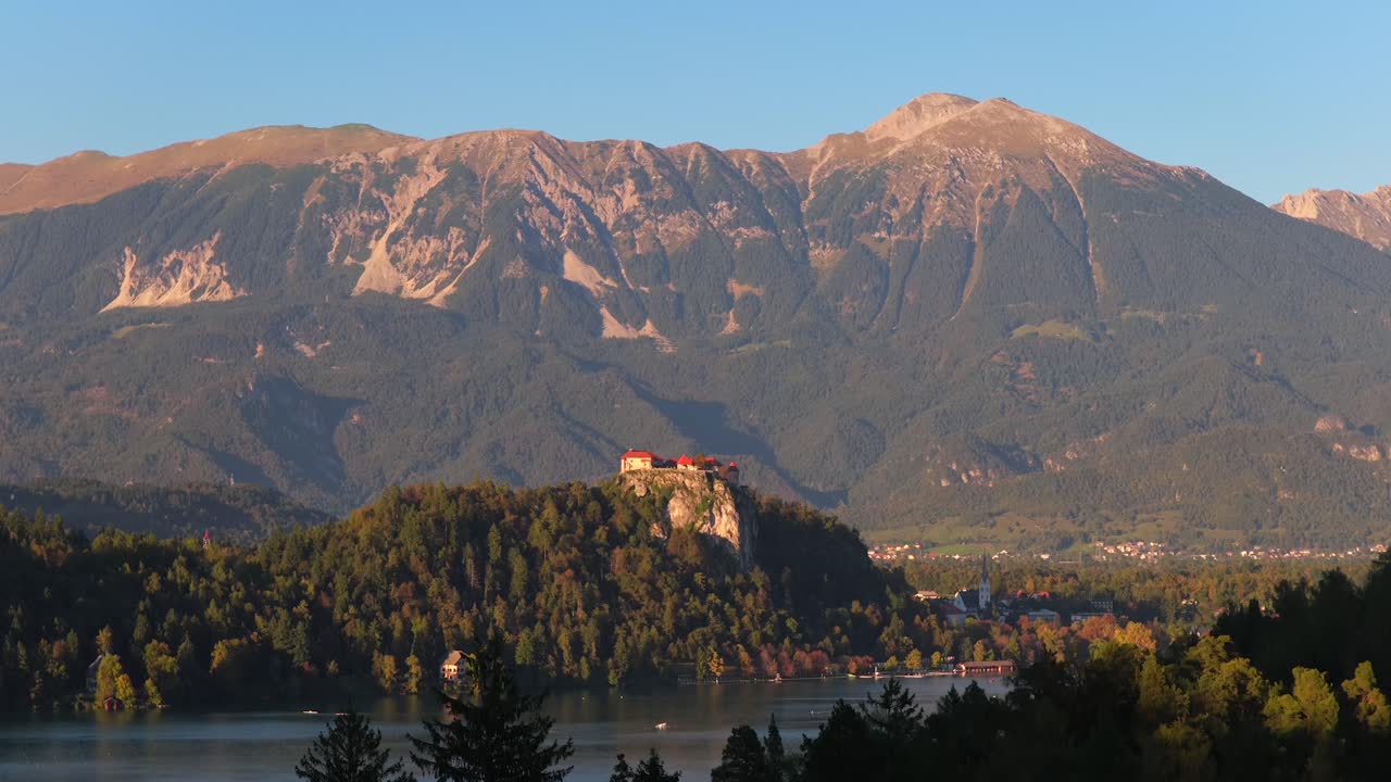 Vista aerea del castello di Bled su una scogliera rocciosa sopra il lago con le Alpi Giulie nella calda luce pomeridiana, ripresa dall'alto sopra la silhouette di un albero
