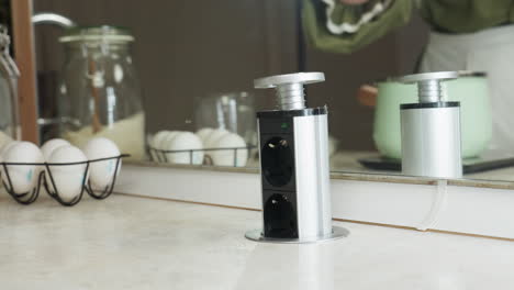 hand view of person pulling up built-in pop-up electrical socket from kitchen counter in preparation to plug in electric twister, with eggs, flour jar, and reflection in background