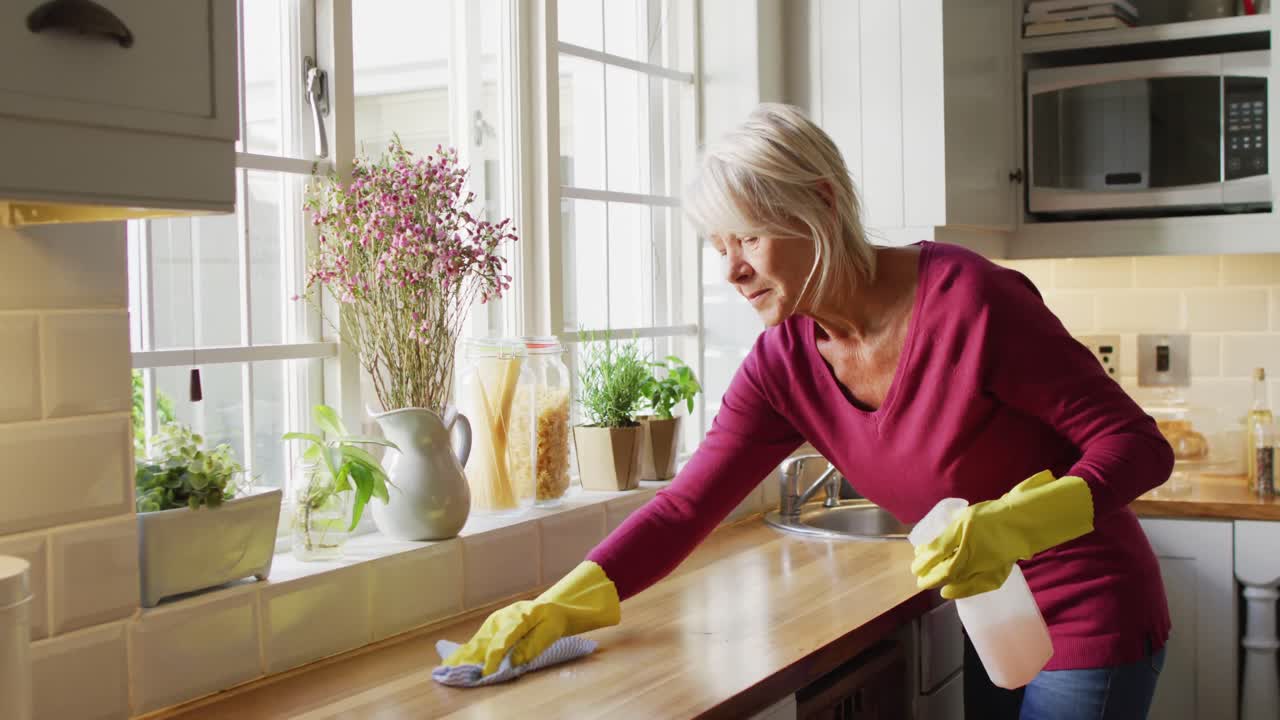 Donna anziana caucasica felice che indossa guanti di gomma e pulisce il piano di lavoro in cucina.