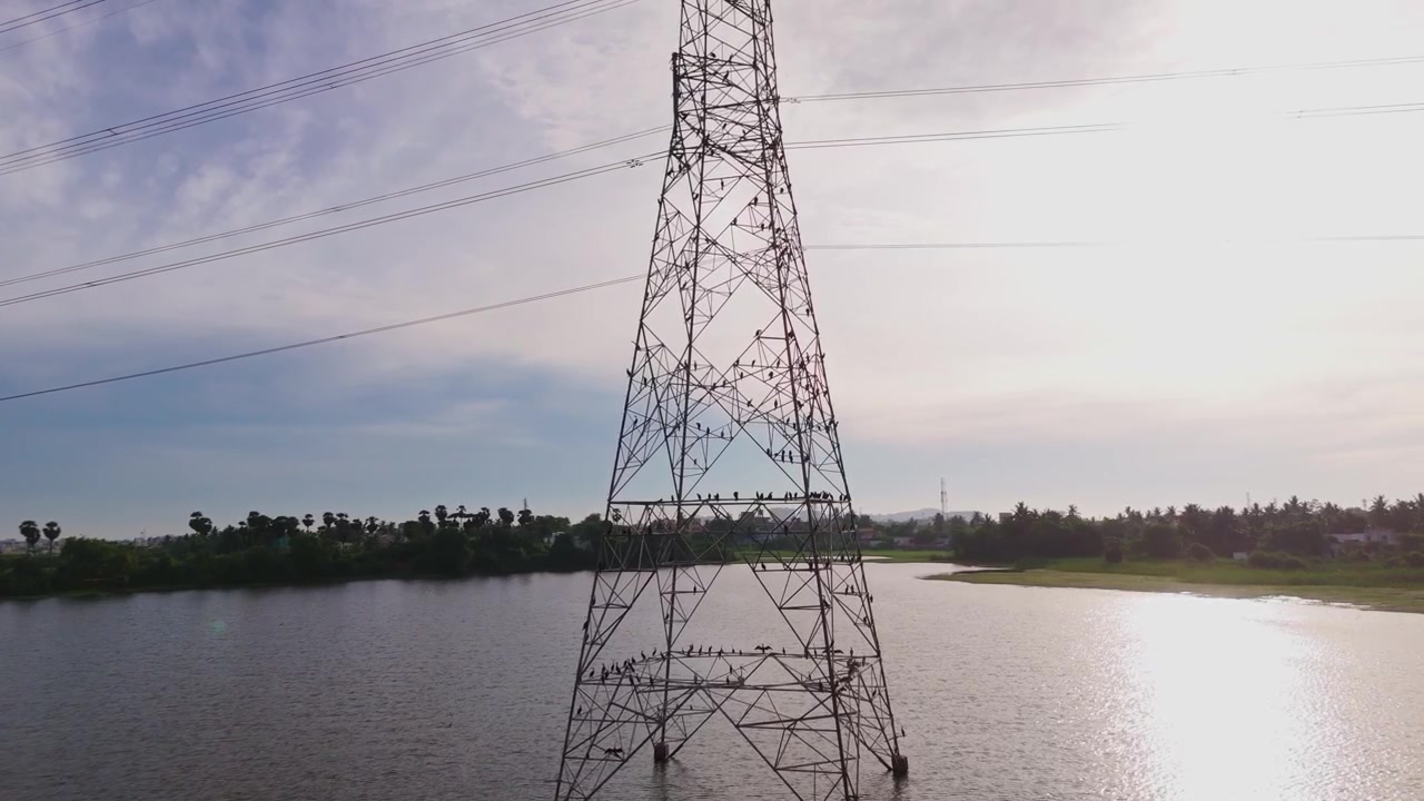Aerial drone shot of power transmission tower standing in lake under bright daytime sky with overhead power lines stretching across calm water and distant green shoreline
