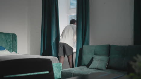 man in white t-shirt standing in bedroom, pulling dark green curtains apart to let sunlight fill room, with green headboard and clean, minimalist bedding and decor