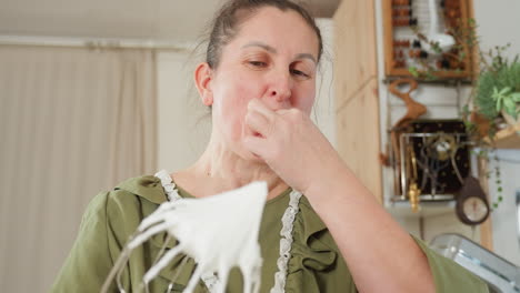 cheerful lady in green apron playfully scoops creamy whipped flour from mixer beater and brings it to her mouth with a joyful smile while baking in cozy kitchen filled with plants and rustic charm