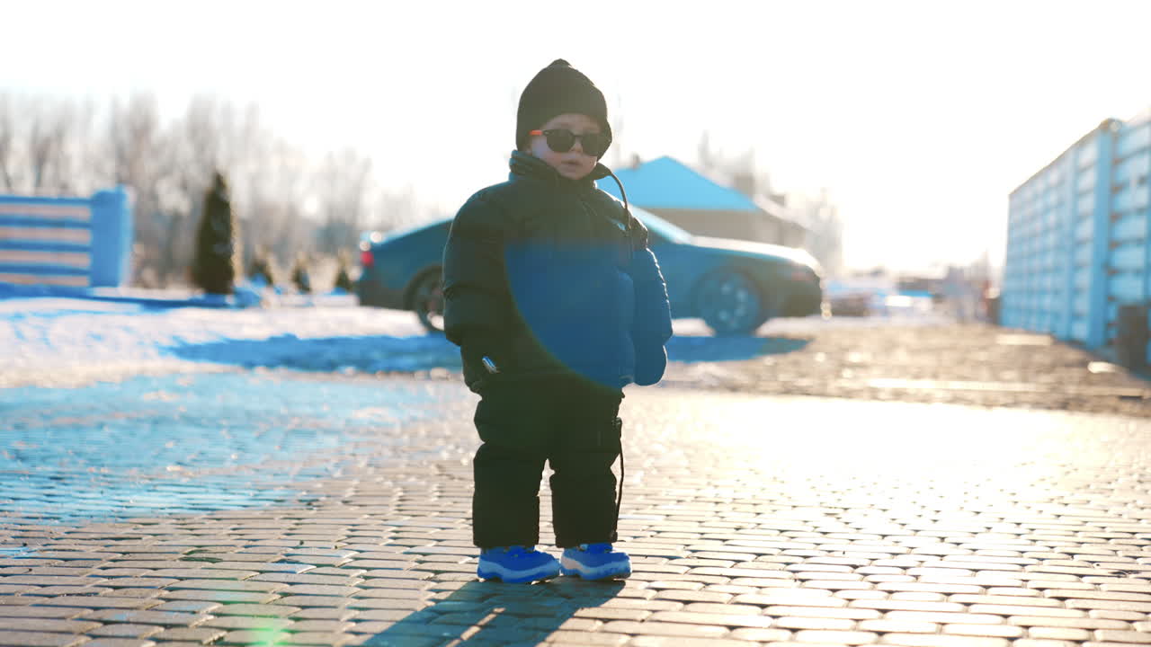 Cute fashionable baby boy wearing black cap, blazer, pants and blue shoes. Kid stands outdoors holding key from car at backdrop.