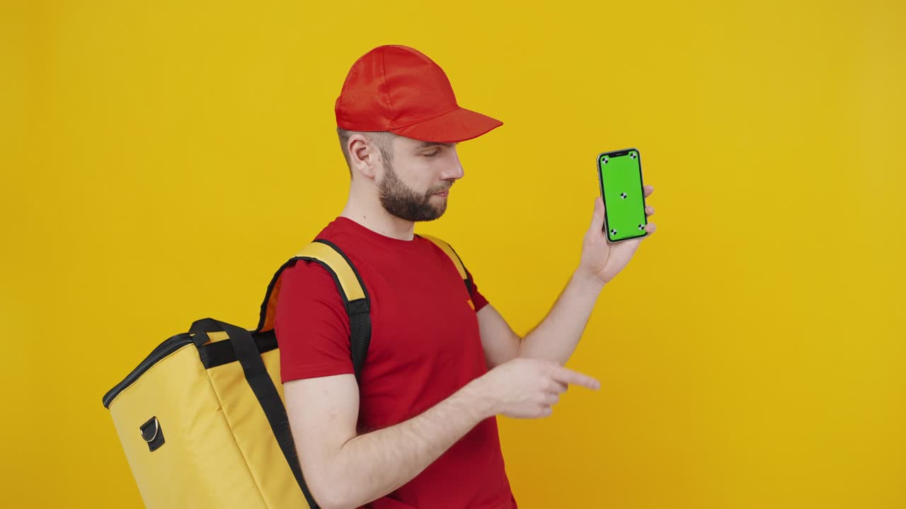 Joven repartidor positivo usando smartphone, aprobando aplicación y mostrando dispositivo con pantalla de croma para maqueta